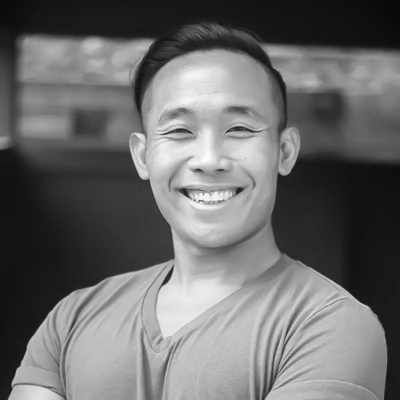 Black and white headshot of Fran Xavier smiling warmly with arms crossed, wearing a casual V-neck t-shirt against a softly blurred background.